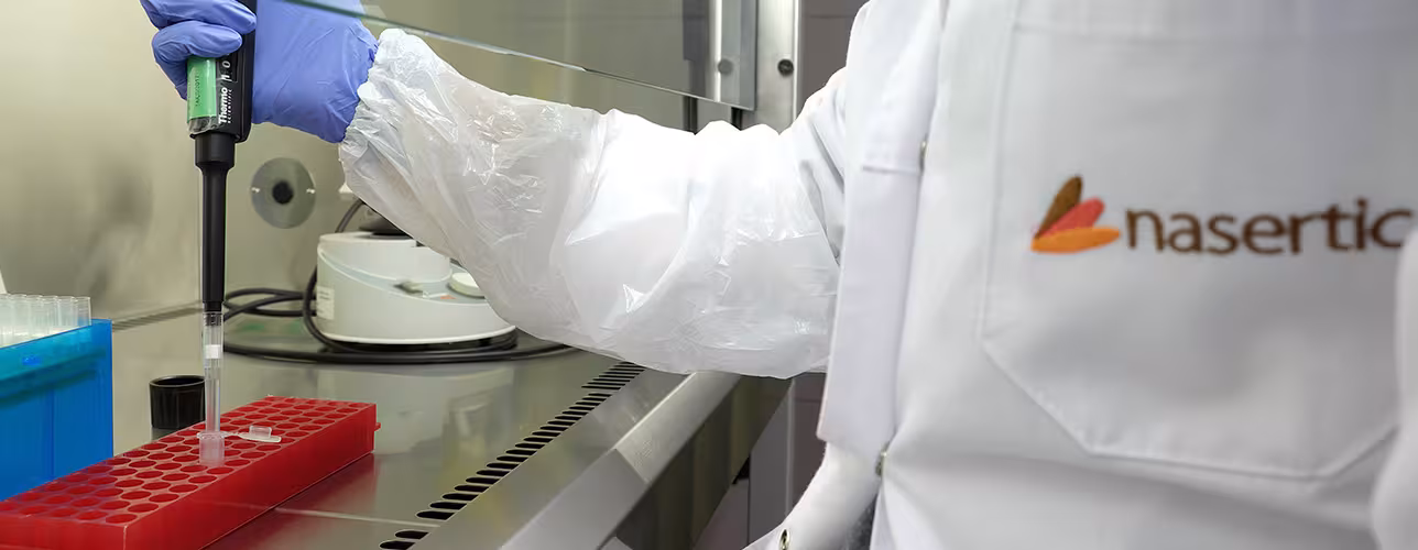 Mano con guante usando una micropipeta para transferir líquido a tubos de ensayo en una bancada de laboratorio; la bata blanca muestra el logotipo de Nasertic.