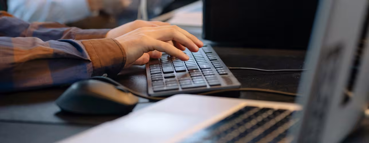Primer plano de una persona escribiendo en un teclado mecánico junto a un ratón y un ordenador portátil sobre un escritorio oscuro.